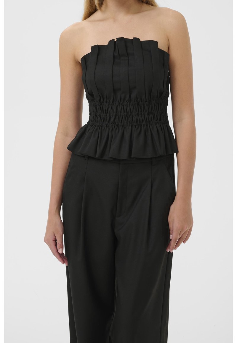 Haut noir sans bretelles avec des détails plissés, taille froncée et ourlet à volants porté avec un pantalon noir ample sur une personne debout.