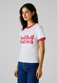 Tricou alb cu guler și mâneci roșii, având logo-ul îndrăzneț "Red Bull Racing" în print roșu pe față. Material din bumbac.