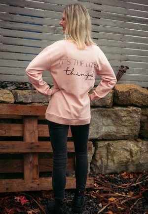 Rosa Sweatshirt mit schwarzem Text "ES SIND DIE KLEINEN DINGE" auf dem Rücken, getragen mit dunklen Jeans und schwarzen Schuhen. Der Text hat eine moderne Schriftart.