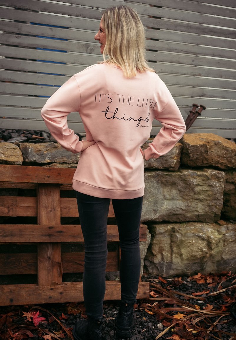 Rosa Sweatshirt mit schwarzem Text "ES SIND DIE KLEINEN DINGE" auf dem Rücken, getragen mit dunklen Jeans und schwarzen Schuhen. Der Text hat eine moderne Schriftart.