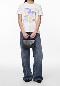 Bílý tričko s barevným geometrickým potiskem v purpurové a žluté, spárované se širokými modrými džínami a černou kabelkou s krokodýlí texturou.