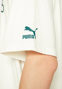 Bílý bavlněný tričko s vytisknutým zeleným logem Puma na levém rukávu. Tkanina je hladká a měkká s uvolněným střihem.