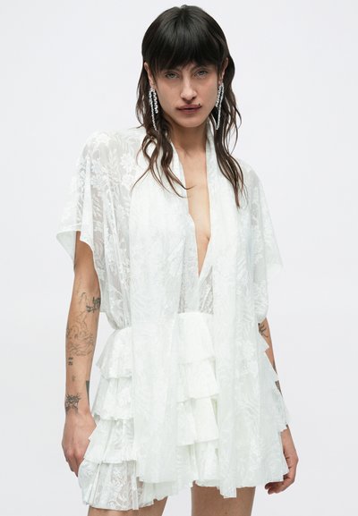 Osoba s tmavými vlasmi a tetovaniami nosí biele čipkované šaty s vrstvenou sukňou a hlbokým výstrihom do V, doplnené dlhými krištáľovými náušnicami.