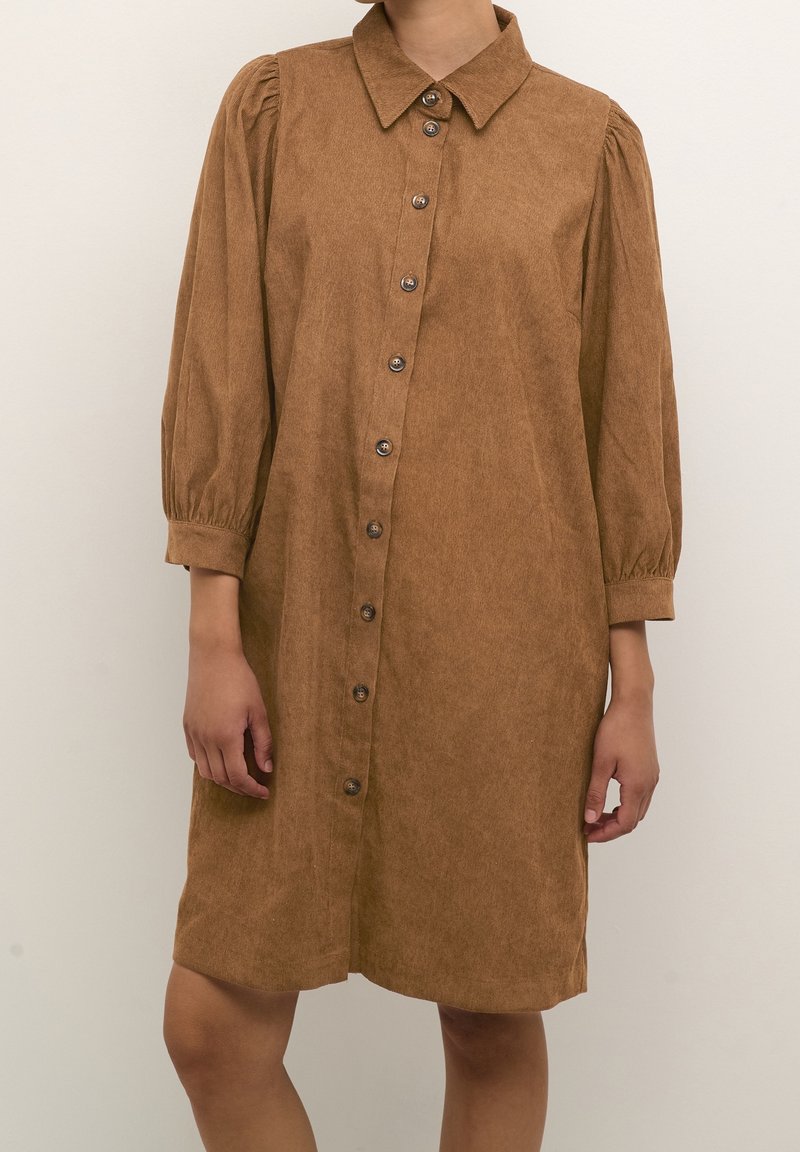Robe en velours côtelé marron à boutons, manches bouffantes trois quarts et col pointu, portée par une personne debout sur un fond uni.