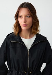 Zwarte ritsjack met capuchon, elastische taille, gouden ritsdetails en een gladde textuur. Draagt over een wit shirt.