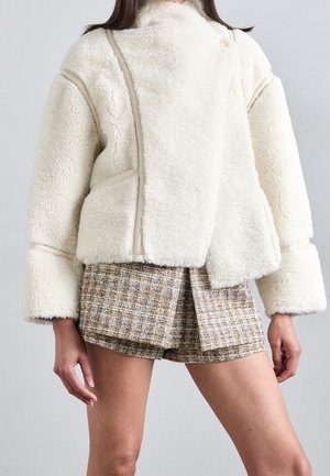 Kvinde iført en hvid faux fleecejakke med brede manchetter og brune ternede omslagsshorts mod en ensfarvet baggrund.