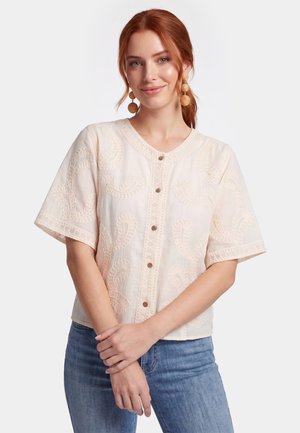 Frau mit roten Haaren, die eine beige bestickte kurzärmelige Bluse mit Knöpfen und blaue Jeans trägt, lächelnd mit vor dem Körper gefalteten Händen.