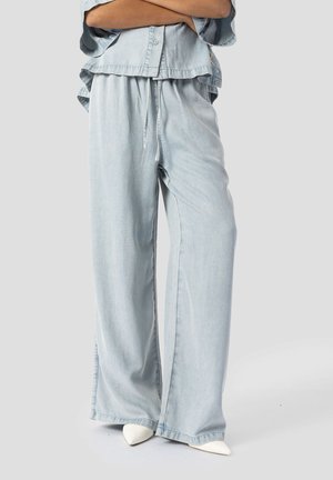 Persona che indossa pantaloni di jeans larghi azzurri con coulisse in vita e camicia azzurra a maniche corte, in piedi con le braccia incrociate.