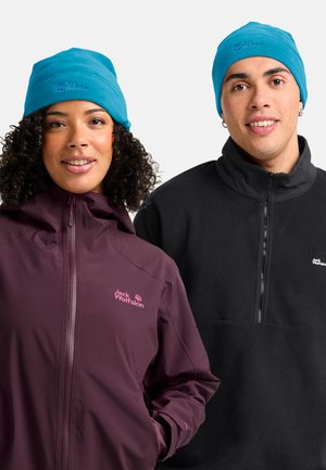 Woman in purple Jack Wolfskin jacket and blue beanie, man in black Jack Wolfskin fleece and blue beanie, both smiling against white background.