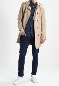 Beige trench coat with double-breasted buttons, navy patterned shirt, navy pants, and white sneakers. Smooth fabric and tailored fit.