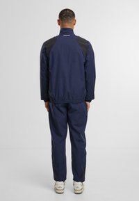 Námořnická větruodolná bunda s černými akcenty na ramenech, elastickým lemem a manžetami, v kombinaci s odpovídajícími námořnickými kalhotami a bílými teniskami.