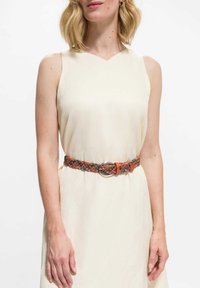 Pleciony pasek z wielokolorowego plecionego materiału z pomarańczowymi akcentami i srebrną metalową klamrą, noszony na jasnokremowej sukience.