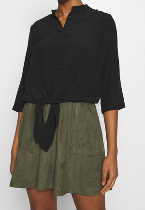 Vrouw draagt een zwarte blouse met lange mouwen en knopen, gestrikt in de taille, en een olijfgroene suède rok met grote voorzakken.