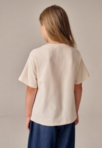 Tricou crem cu mânecă scurtă, fabricat din bumbac; are o croială lejeră și un guler rotund. Asortat cu pantaloni din denim bleu închis.