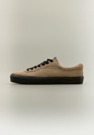 Baskets basses beiges en suède avec semelle en caoutchouc noir et lacets noirs, présentées de profil sur fond neutre.