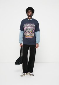 Tricou grafic bleumarin cu imprimeul "CROWN JEWEL KINGS LOS ANGELES", cu mâneci de un albastru deschis, asortat cu pantaloni largi negri și adidași.