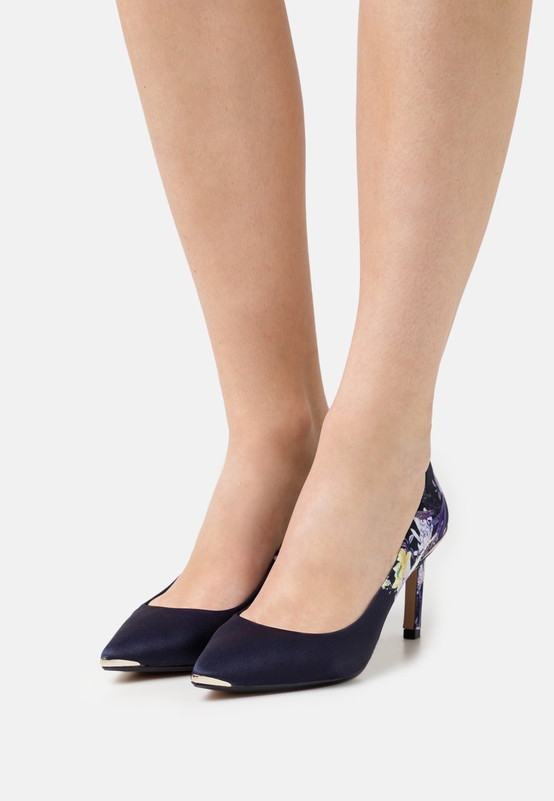 Femme portant des escarpins bleu marine à bout pointu avec talons à motif floral, debout sur un fond blanc.