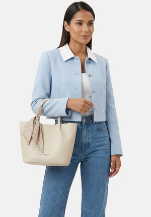 Nainen pukeutunut vaaleansiniseen cropattu takkiin ja farkkuihin, pitää kädessään beigeä käsilaukkua, jossa on kuviollinen huivi sidottuna kahvaan.