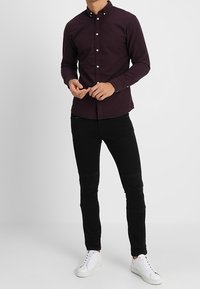 Muž oblečený v tmavě pruhované košili na knoflíky, černých slim-fit džínách a bílých teniskách, stojící před jednobarevným světlým pozadím.