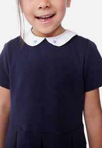 Ναυτικό μπλε φόρεμα με κοντά μανίκια, που διαθέτει λευκό γιακά κεντημένο με δύο μπλε καρδιές. Η φούστα έχει πτυχές για επιπλέον λεπτομέρεια.