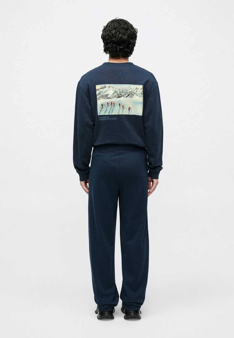 Ensemble sweat à capuche et pantalon bleu marine foncé, en tissu doux, avec une image en couleur au dos représentant des skieurs dans un paysage enneigé. Coupe décontractée, poignets côtelés.