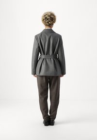 Sivý vlnený blazer s opaskom v oblasti pása a skladaným zadným dielom, nosený cez hnedé textúrované nohavice. Model má krátke, kučeravé vlasy.