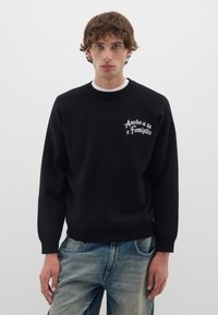 Felpa nera con collo a giro e maniche lunghe, caratterizzata da testo ricamato bianco sul lato sinistro del petto. Abbinata a jeans in denim azzurri.