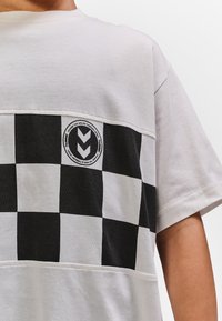 White T-shirt with black and white checkerboard stripe across chest and a round logo with arrows and text on left side.