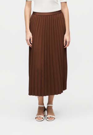 Naine kannab pruuni volditud midi seelikut, mida on kombineeritud valge topi ja valgete avatud varbaga plokktaldsete sandaalidega, millel on pahkluupiirded.