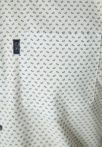 Camisa de manga corta en tela ligera con fondo blanco, que presenta un patrón floral en azul marino. Incluye un bolsillo en el pecho izquierdo y una etiqueta con el logo.