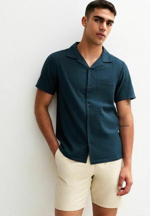 Chemise à manches courtes en teal foncé avec un tissu texturé, col à revers et une poche poitrine, associée à un short beige clair.