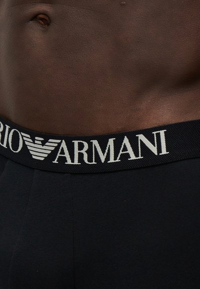 Gros plan sur un torse à la peau foncée portant un sous-vêtement noir avec une ceinture noire portant le logo blanc Emporio Armani et le symbole de l'aigle.