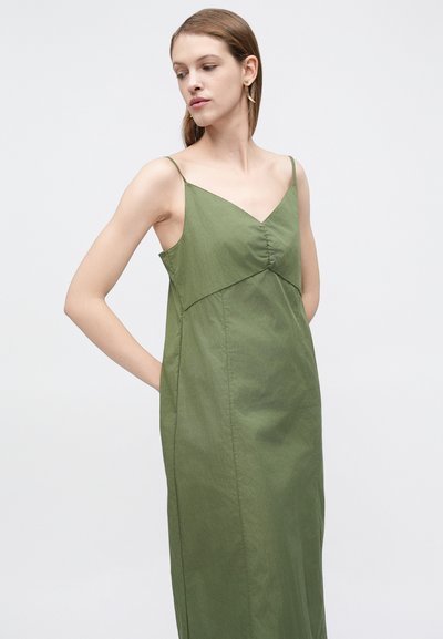 Γυναίκα με αμάνικο ελιάς πράσινο φόρεμα με λεπτές τιράντες, τα χέρια πίσω από την πλάτη, κοιτάζοντας πλάγια σε απλό φωτεινό φόντο.