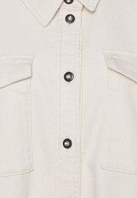 Biała koszula dżinsowa z wyraźnym kołnierzykiem, dwiema kieszeniami na piersi oraz czarnymi guzikami z przodu. Teksturowany materiał o subtelnej strukturze.