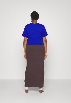 Vrouw staat met haar rug naar de camera, draagt een blauw kortemouwshirt, een lange bruine rok en witte hakken tegen een lichte effen achtergrond.