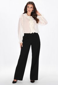 Norsunluunvalkoinen paita, jossa liehutetut reunat ja solmittava kauluspanta, yhdistetty korkeavyötäröisiin mustiin leveälahkeisiin housuihin ja mustiin korkokenkiin.