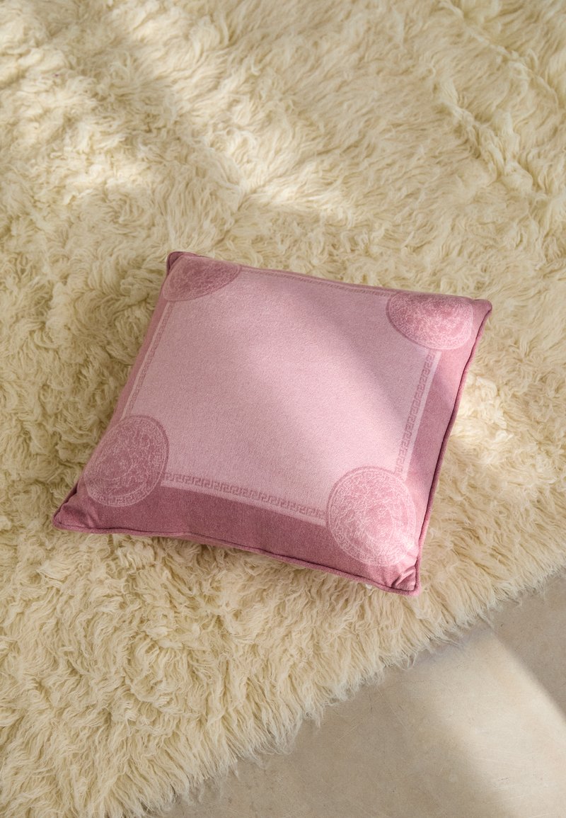 Square pink cushion made of soft velvet, featuring a subtle border design and round accent details at each corner, placed on a textured surface.