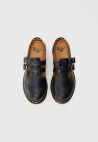 Chaussures Mary Jane Dr. Martens en cuir noir avec double bride à boucles, motif cœur perforé sur l’avant et coutures jaunes sur la semelle.