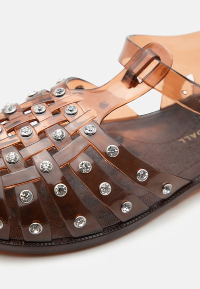 Καφέ διαφανές σανδάλι με σταυρωτούς ιμάντες διακοσμημένους με ασημένιες στρογγυλές καρφίτσες και κλείσιμο με αγκράφα στον αστράγαλο.