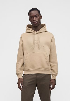 Homme portant un sweat à capuche beige avec poche avant et "Originals" brodé sur la poitrine, associé à un pantalon marron foncé.