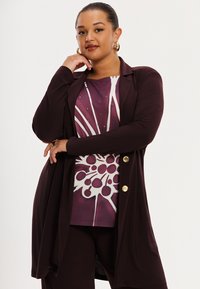 Maroon long cardigan over a patterned purple top with white accents. Features large buttons and soft fabric. Smooth texture, modern design.