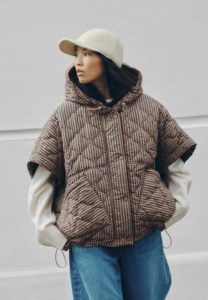 Femme portant une veste matelassée à manches courtes rayée marron, un pull crème, un bonnet en tricot beige et un jean bleu, debout contre un mur blanc.