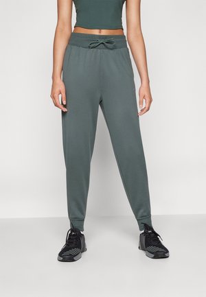 Kobieta ubrana w ciemnozielone spodnie dresowe ze ściągaczem w pasie i czarne buty sportowe, stojąca na tle jednolitego jasnego tła.