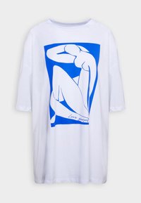 Camiseta blanca de gran tamaño con una ilustración gráfica azul de una figura sentada, con el texto "Ámate a ti mismo" integrado en la parte inferior.