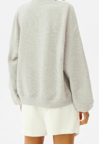 Šedá příliš velká mikina s žebrovaným límcem a manžetami, vyrobená z měkkého materiálu, spojená s bílými šortkami. Má uvolněný střih.