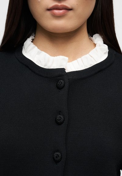 Κοντινό πλάνο ενός ατόμου που φοράει μαύρο πλεκτό ζακέτα με κουμπιά καλυμμένα με ύφασμα πάνω από μια λευκή μπλούζα με βολάν στο γιακά.