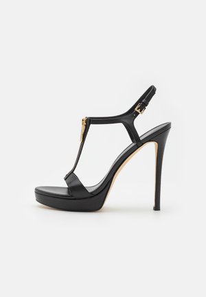 Sandale à plateforme noire à talons hauts avec fine bride à la cheville, détail fermeture éclair à l'avant et boucle dorée sur fond blanc.