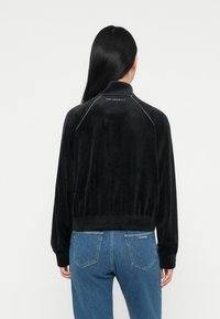 Fekete velúr dzseki magas gallérral, bordázott aljjal és fehér díszvarrással. A logó a hátsó részen látható, kék farmernadrággal viselve.