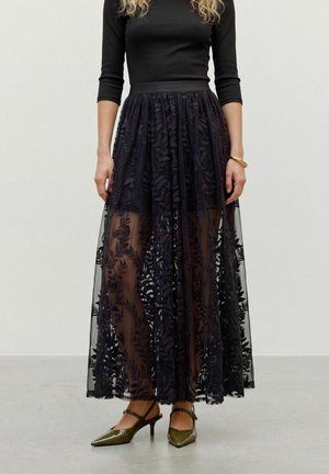 Un mannequin porte un haut noir à manches longues, une jupe longue transparente à motif floral, un bracelet jonc doré et des escarpins pointus couleur olive.