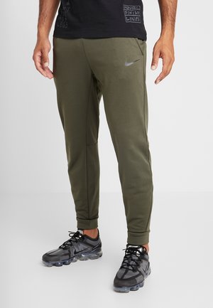 Mann trägt olivgrüne Nike-Jogginghosen und schwarze Nike-Sneaker, steht entspannt mit den Händen an den Seiten vor einem schlichten Hintergrund.